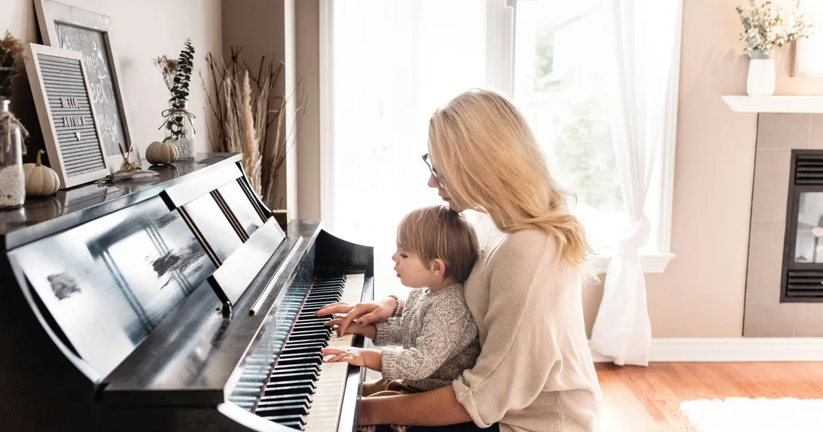 A mother and her young child sitting together at an upright piano, playing the keys