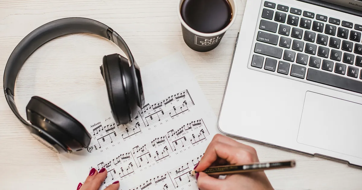 An adult’s hands annotating sheet music with a pencil, beside headphones, a coffee and a laptop